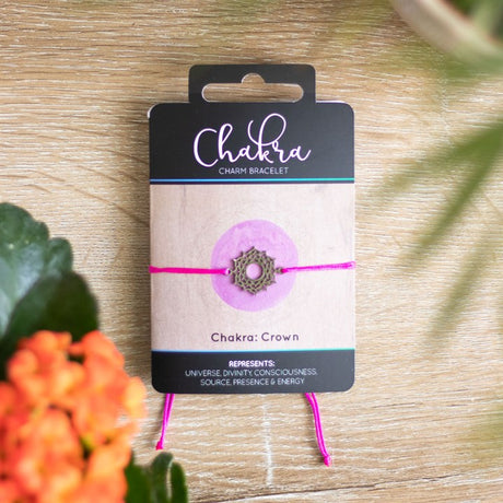 A Crown Chakra charm bracelet with a pink band and a silver charm, displayed on a card with 'Chakra' written on it, placed on a wooden surface with orange flowers in the background.