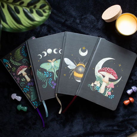 A set of A5 notebooks with a dark forest theme, featuring illustrations of mushrooms, a luna moth, and other mystical plants. The notebooks have gold foil page edges and a ribbon marker.