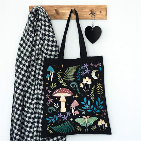 A black cotton tote bag with a mystical dark forest mushroom design, hanging on a wooden hook next to a houndstooth patterned jacket and decorative black heart