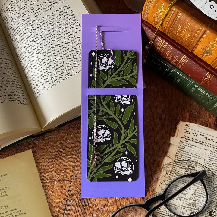 Purple card with black and green bookmark skull and plant design on a wooden surface with books and glasses.