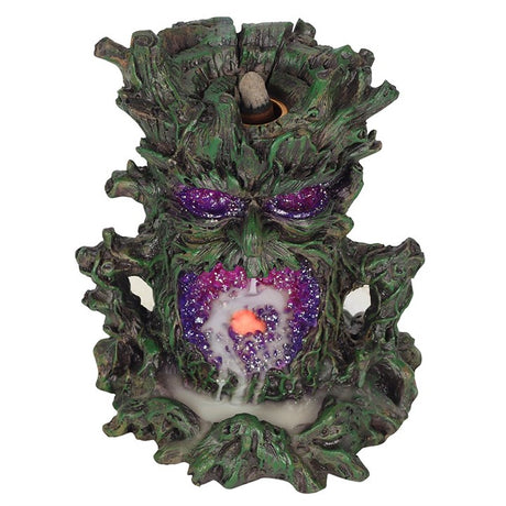 A dark-coloured back flow incense burner designed to resemble a stylised green man face with leaf-like patterns, featuring a lighted crystal cave for smoke to flow through, set against a white backdrop.