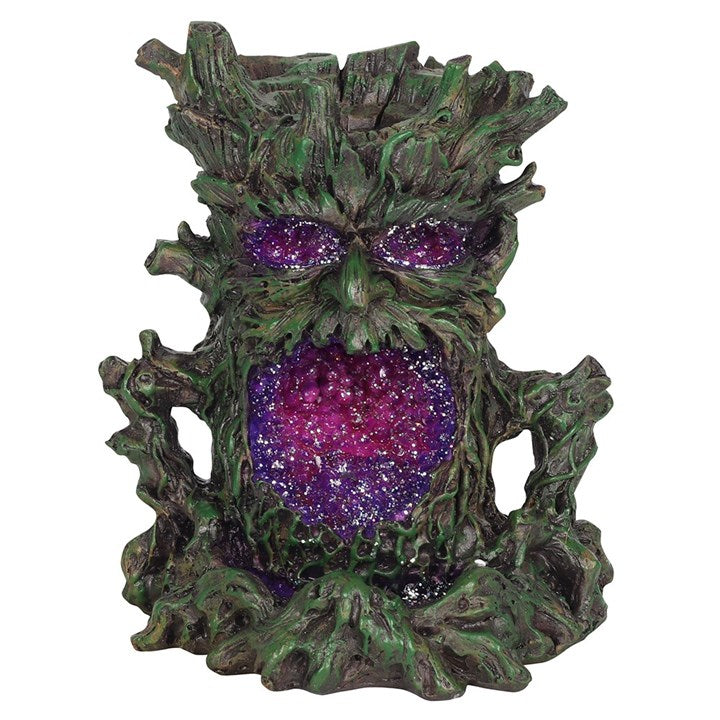 A dark-coloured back flow incense burner designed to resemble a stylised green man face with leaf-like patterns, featuring a lighted crystal cave for smoke to flow through, set against a white backdrop.