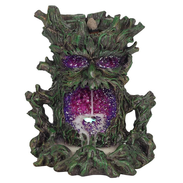 A dark-coloured back flow incense burner designed to resemble a stylised green man face with leaf-like patterns, featuring a lighted crystal cave for smoke to flow through, set against a white backdrop.