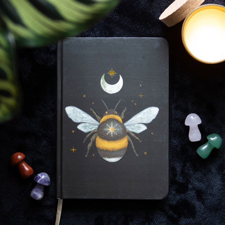 A dark forest bee and moon themed A5 black notebook on a dark surface surrounded by crystal mushrooms. 