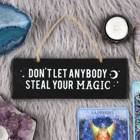 A black hanging wall sign with white text that reads 'DON'T LET ANYBODY STEAL YOUR MAGIC', accompanied by a crescent moon symbol. The sign is displayed on a grey, furry surface, with a candle, crystal, and tarot card arranged around it.