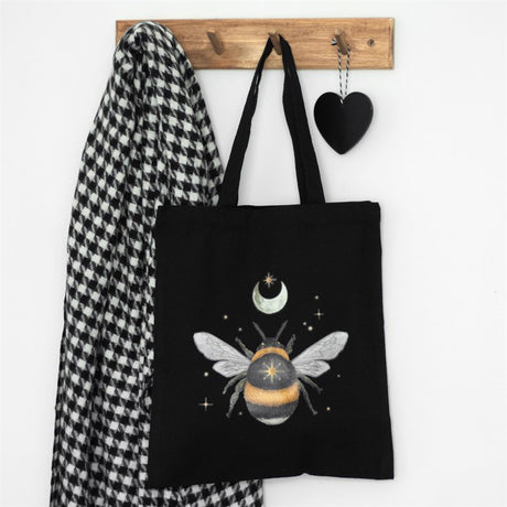 A black cotton tote bag with a forest bee print, displayed next to a checkered scarf on a wall hook.