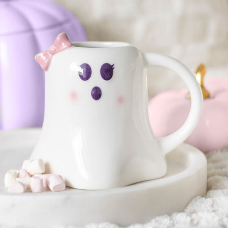 A white ceramic ghost-shaped mug with a pink bow design, displayed on a marble surface with a few marshmallows in front.
