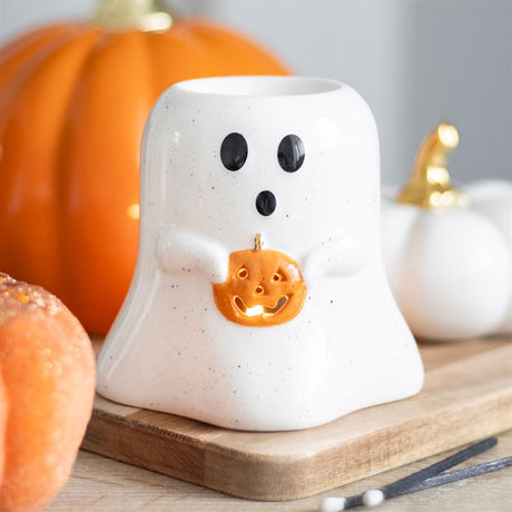 A white ghost-shaped oil burner with a pumpkin design, standing on a wooden surface with pumpkins in the background.