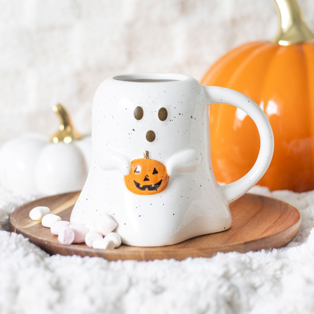 A white ceramic ghost-shaped mug holding a pumpkin, displayed on a wooden surface with a few marshmallows in front.