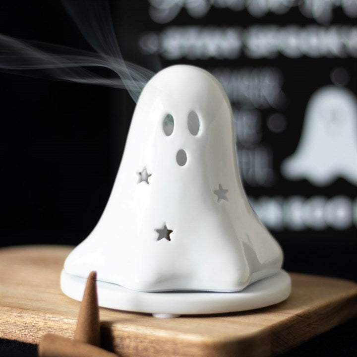 A white ceramic tealight and incense cone holder in the shape of a ghost with a lit incense cone inside, placed on a wooden board.
