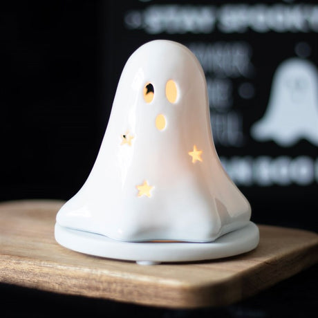 A white ceramic tealight and incense cone holder in the shape of a ghost with a lit tealight inside, placed on a wooden board.