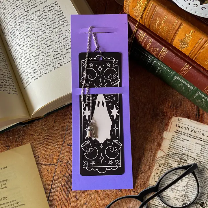 Purple packaging holding a bookmark with ghost design on a wooden surface with books and glasses.