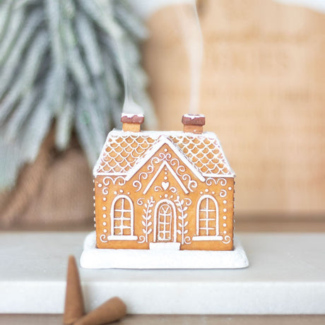 A gingerbread house-shaped incense cone burner with a white base and decorative details, with an incense cone placed under it and smoke rising from the top.