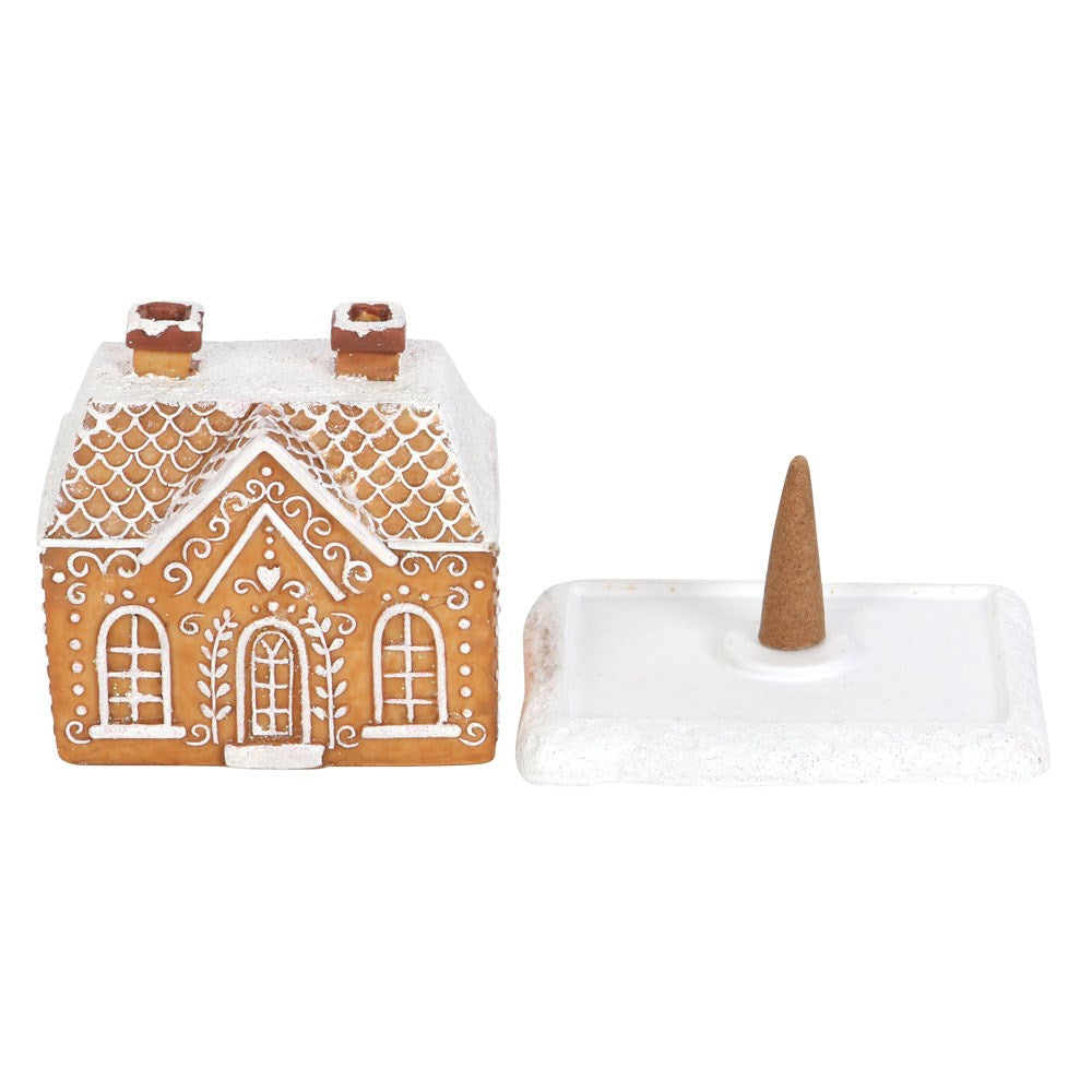 A gingerbread house-shaped incense cone burner with a white base and decorative details, shown with an incense cone placed on it to the side 