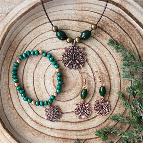 A pair of green man themed green glass bead earrings with bronze-coloured stainless steel hooks, displayed on a wooden surface next to other green man themed jewellery such as a necklace and bracelet