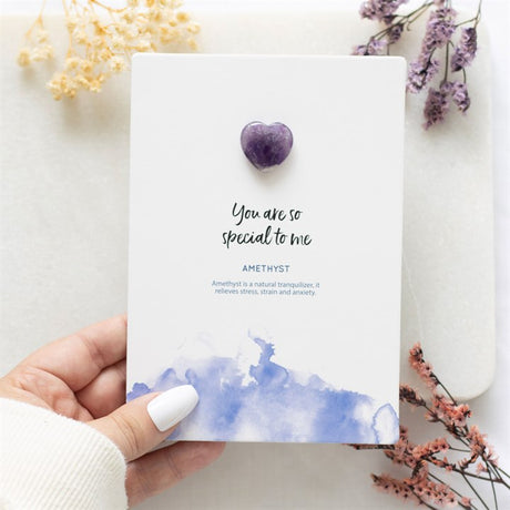 A hand holding a greeting card with a heart-shaped Amethyst crystal attached to it, placed against a floral backdrop.