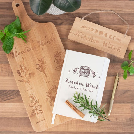 Image shows a "healing herbs" chopping board with etched herb designs, a white "kitchen witch spells & recipes" notebook, a pen and a "kitchen witch" rectangular sign all laid on a wooden background with some herbs scattered. 