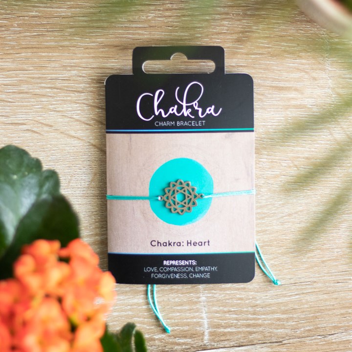 A Heart Chakra charm bracelet with a greenish band and a silver charm, displayed on a card with 'Chakra' written on it, placed on a wooden surface with orange flowers in the background.