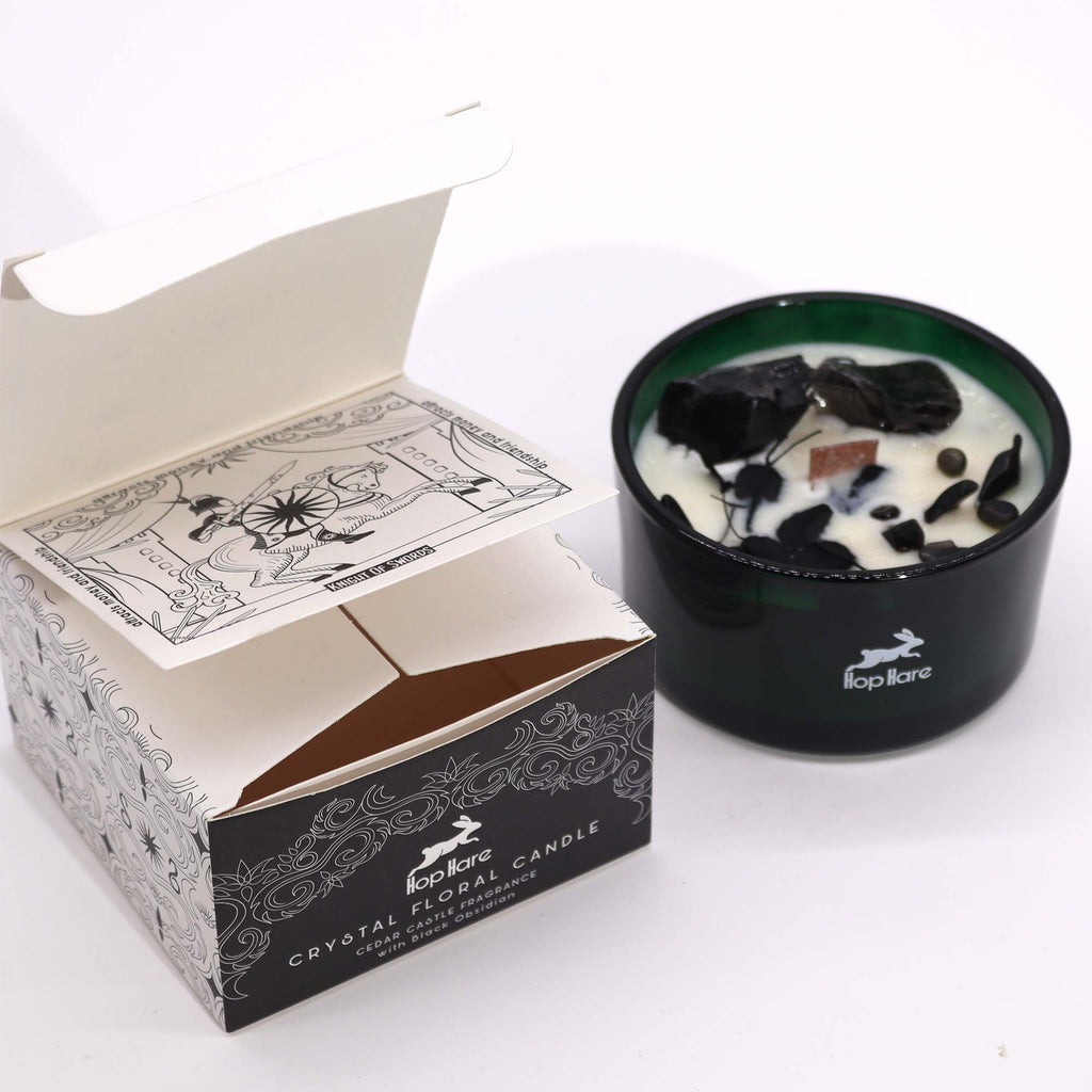 Viewed from the side; a green glass candle with black obsidian gemstones embedded in it and some flowers. Next to it is its matching black box with white detailing. 