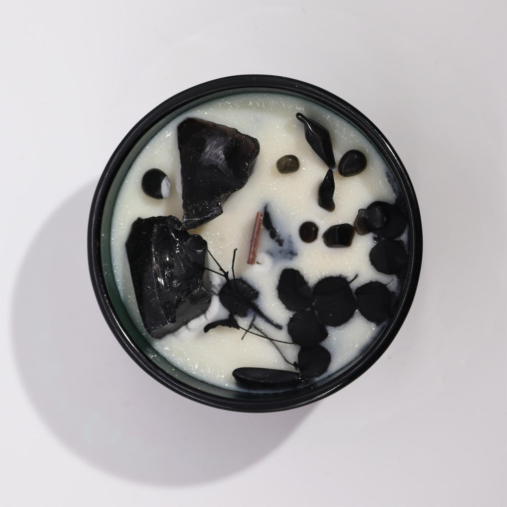 Viewed from above; a green glass candle with black obsidian gemstones embedded in it and some flowers.