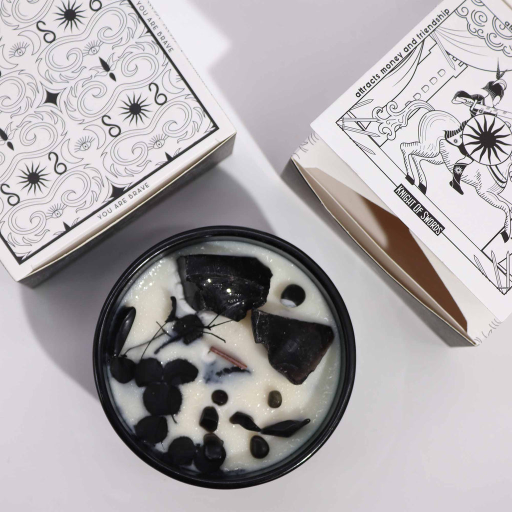 Viewed from above; a green glass candle with black obsidian gemstones embedded in it and some flowers. Above and to the side are two matching white and black boxes with matching detailing. 