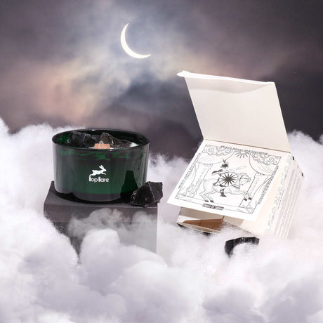 A green glass candle with black obsidian gemstones embedded in it, seeming to float above some clouds and has a tarot card next to it, against a backdrop of a crescent moon in the sky.