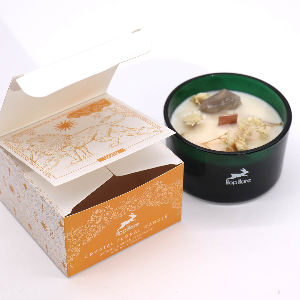 Viewed from the side; a green glass candle with Rock Quartz gemstones embedded in and some dried flowers. To the side, is its matching white and yellow box. 