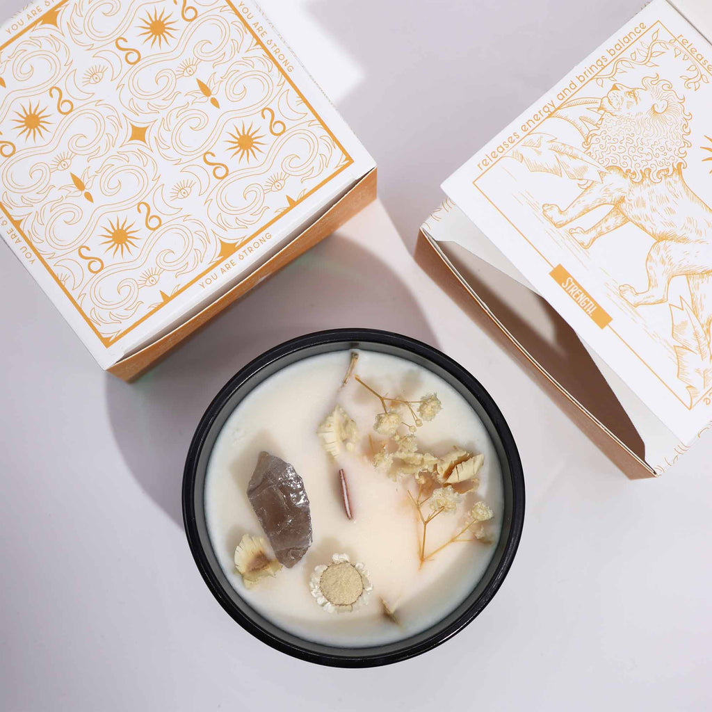 Viewed from above; a green glass candle with Rock Quartz gemstones embedded in and some dried flowers. Above and to the side are two matching white and yellow boxes.