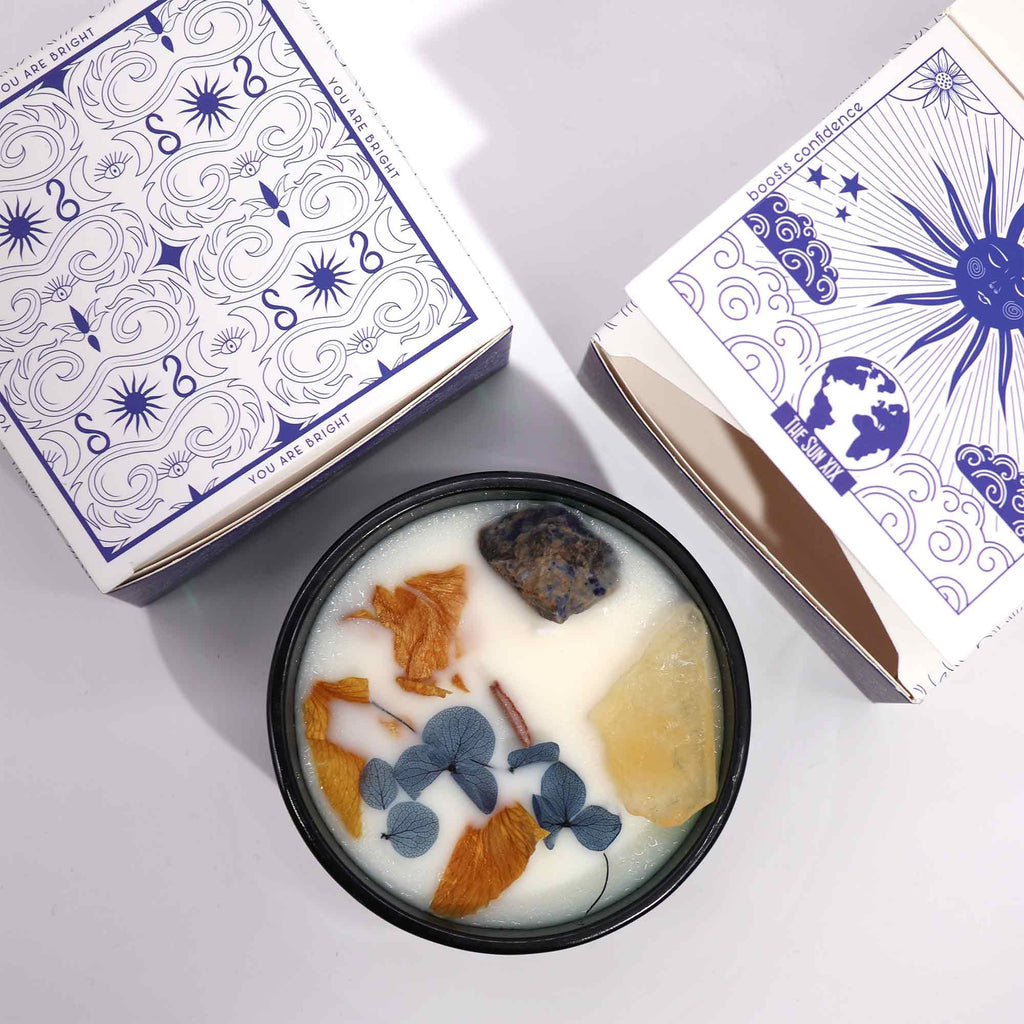 Viewed from above: a green glass candle with natural flowers, blue and yellow gemstones embedded in it.
above and to the side are two matching blue and white boxes. 