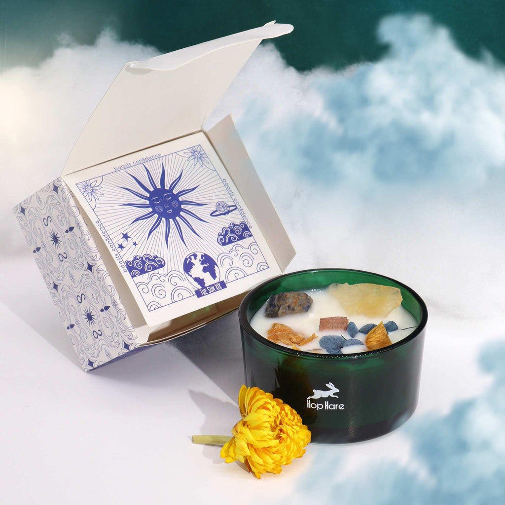 A green glass candle with natural flowers, blue and yellow gemstones embedded in it, presented on a blue fabric surface with a yellow flower decoration beside it.
Above and to the side is its matching box which is white and blue.