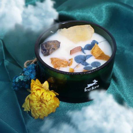 A green glass candle with natural flowers, blue and yellow gemstones embedded in it, presented on a blue fabric surface with a yellow flower decoration beside it.