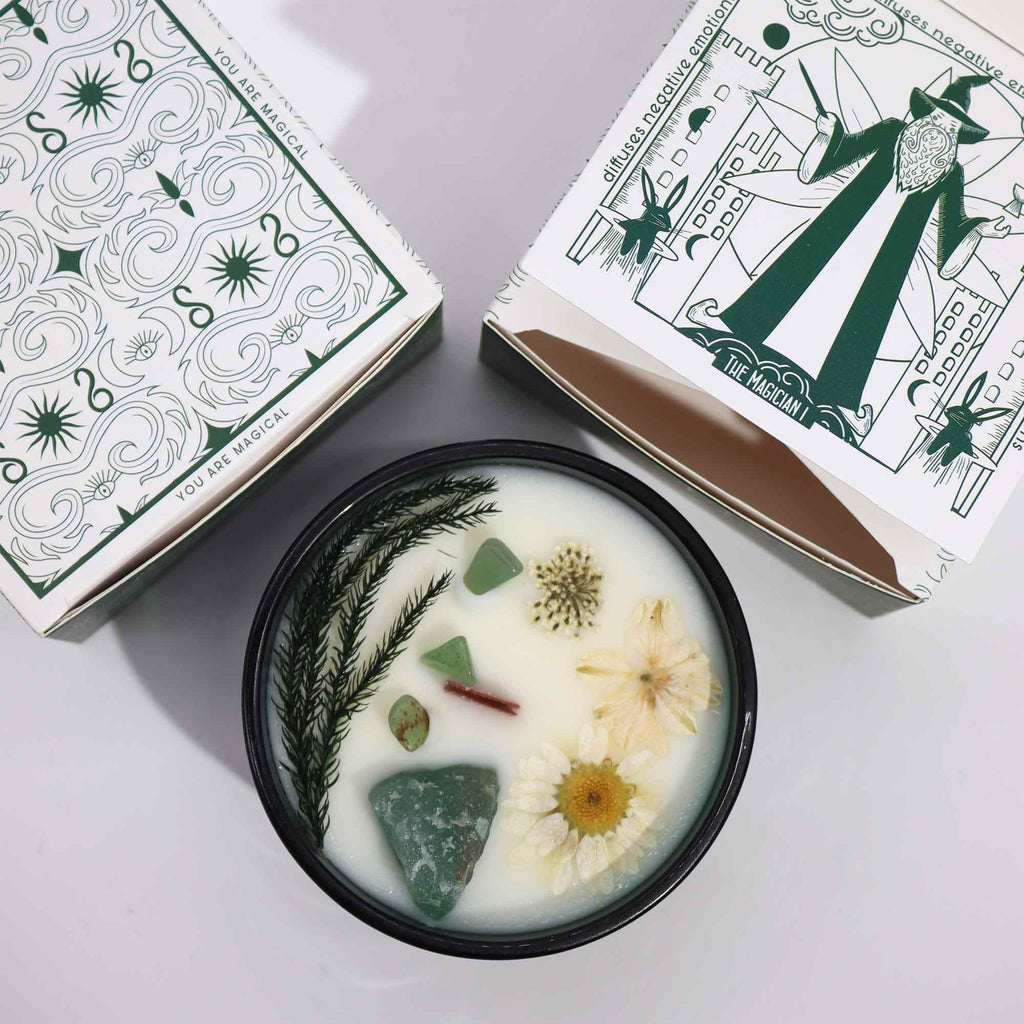 Viewed from above: A green glass crystal magic flower candle, adorned with dry flowers and jade gemstones. There are also two matching green and white boxes including a tarot design.
