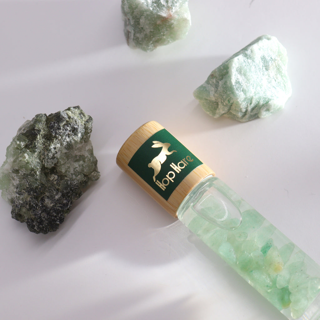 A product image featuring a roll-on essential oil bottle with gemstone chips inside, alongside raw green aventurine stones on a white background.