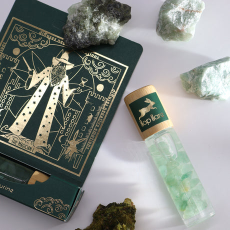 A product image featuring a roll-on essential oil bottle with gemstone chips inside, alongside a green box with gold-printed tarot cards and raw green aventurine stones.