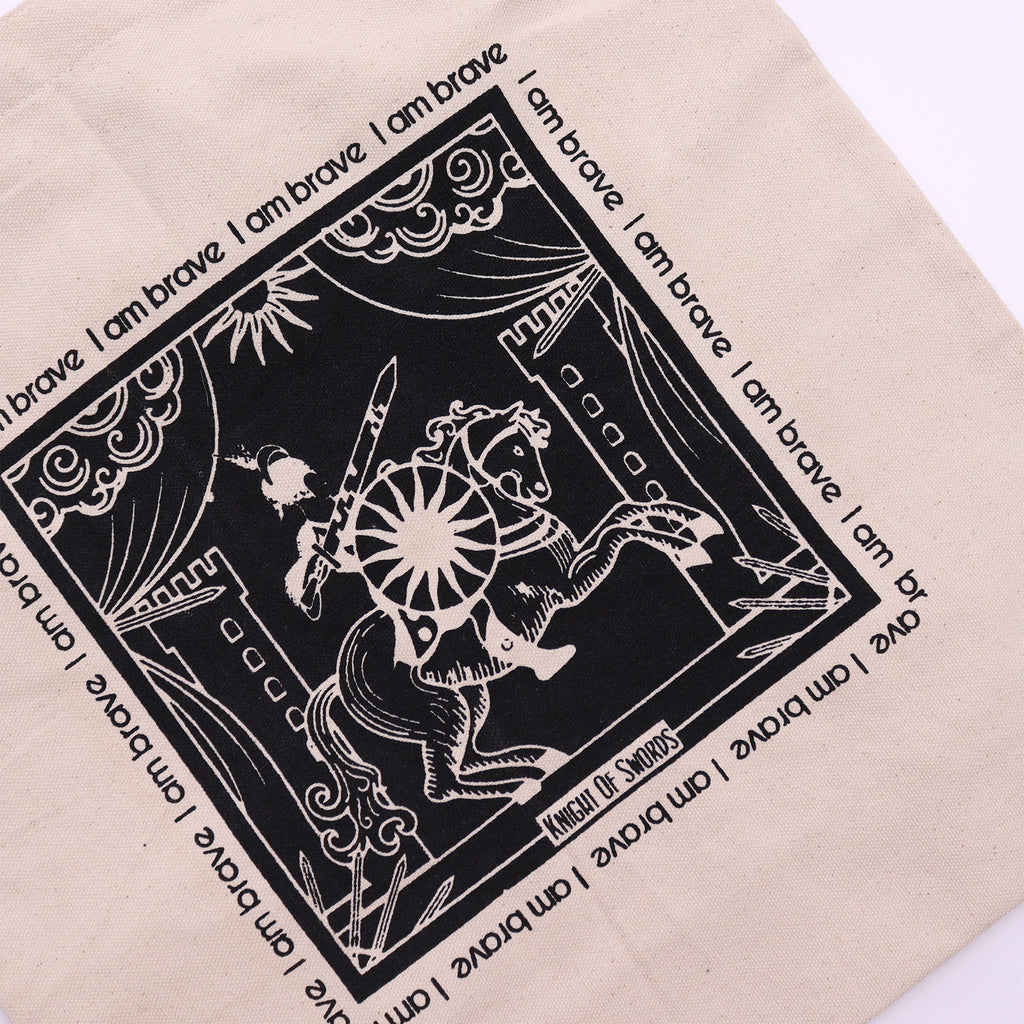 Tote bag with black motif which is of a "knight on a horse" standing in front of a castle with the affirmation "I am brave" repeated around the motif. 