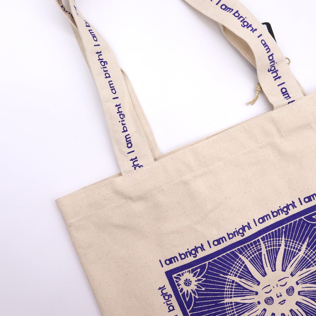 Tote bag with purple motif which is of a sun standing with the affirmation "I am bright" repeated around the motif. 