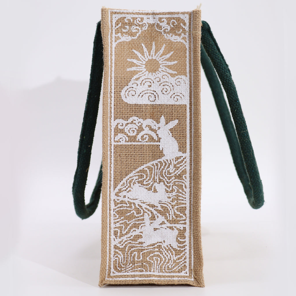 A beige jute bag, with green handles, printed on the side, featuring a moon and hare design.