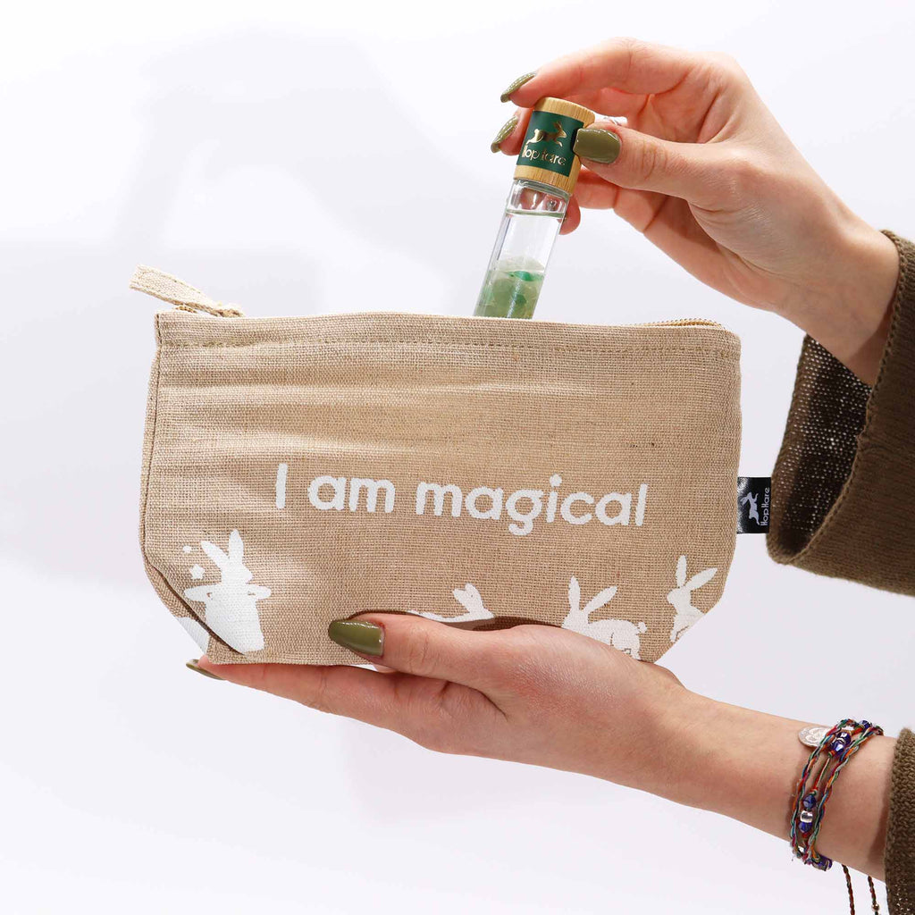 A beige jute pouch with an 'I am magical' affirmation in white, with a person placing an essential oil roll on inside it.