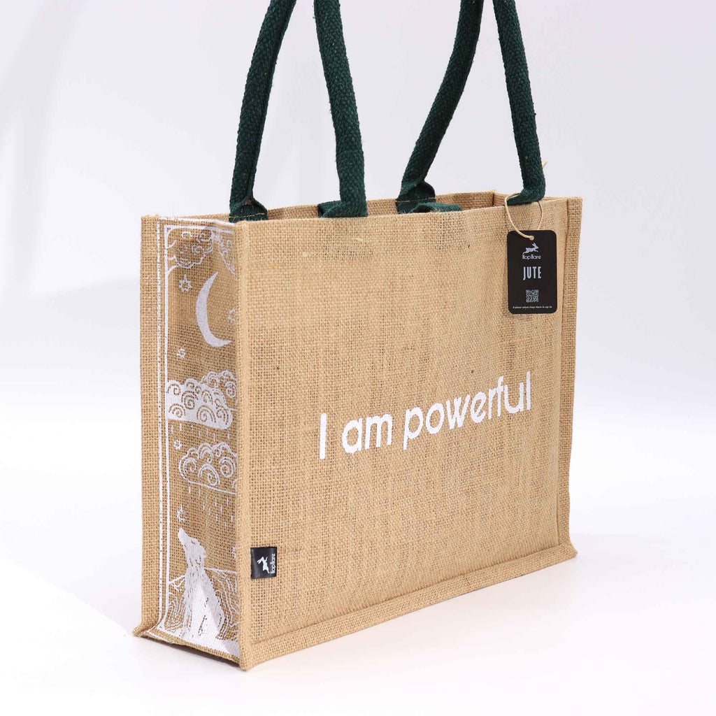 A beige jute bag, with green handles, with the text 'I am powerful' printed on the side, featuring a moon and wolf design.