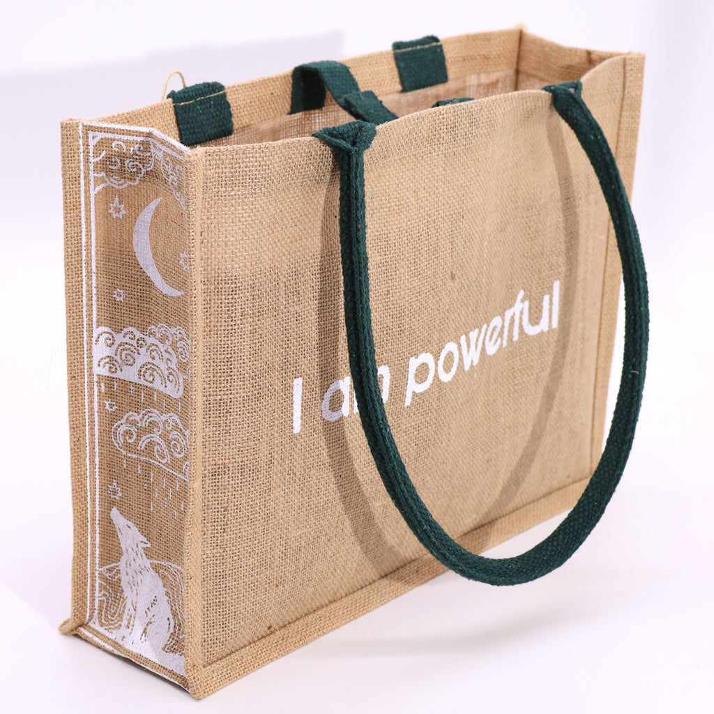 A beige jute bag, with green handles, with the text 'I am powerful' printed on the side, featuring a moon and wolf design.