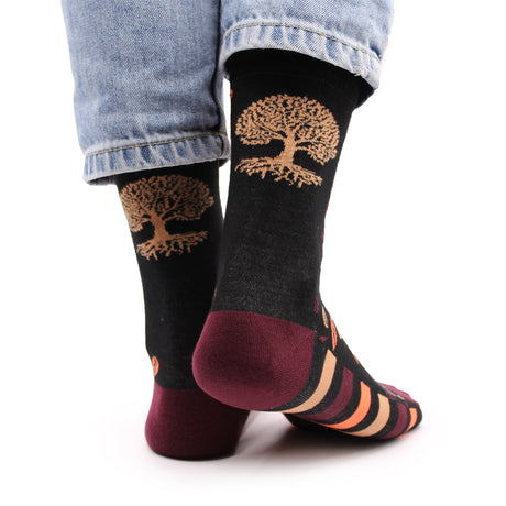 A pair of mostly black bamboo socks with a gold tree of life symbol design, with maroon, cream and pale orange stripes across the bottom and the same coloured leaves across the top, worn on the feet against a white background.