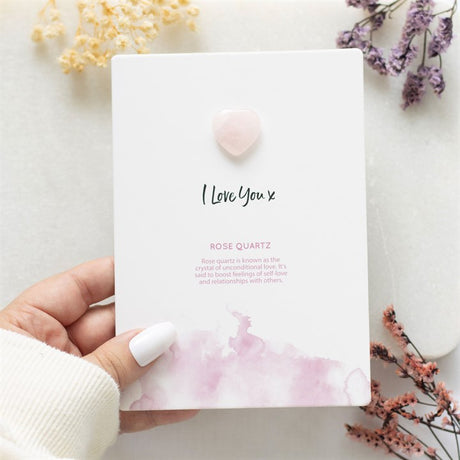 A hand holding a white greeting card with 'I Love You x' text and a small pink Rose Quartz crystal heart attached to it. The card is displayed with some dried flowers around it.