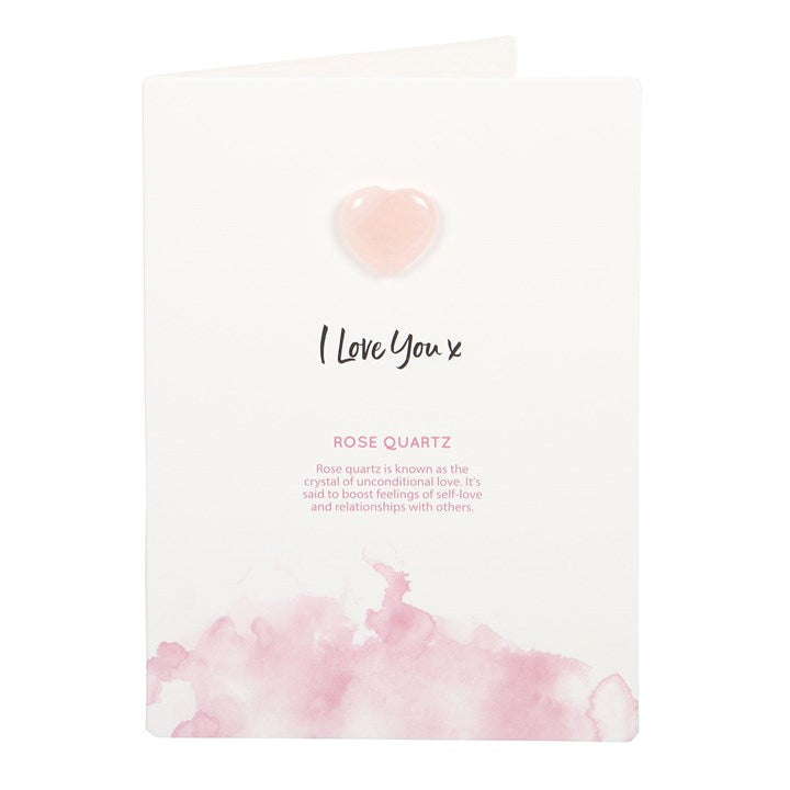 A hand holding a white greeting card with 'I Love You x' text and a small pink Rose Quartz crystal heart attached to it. 