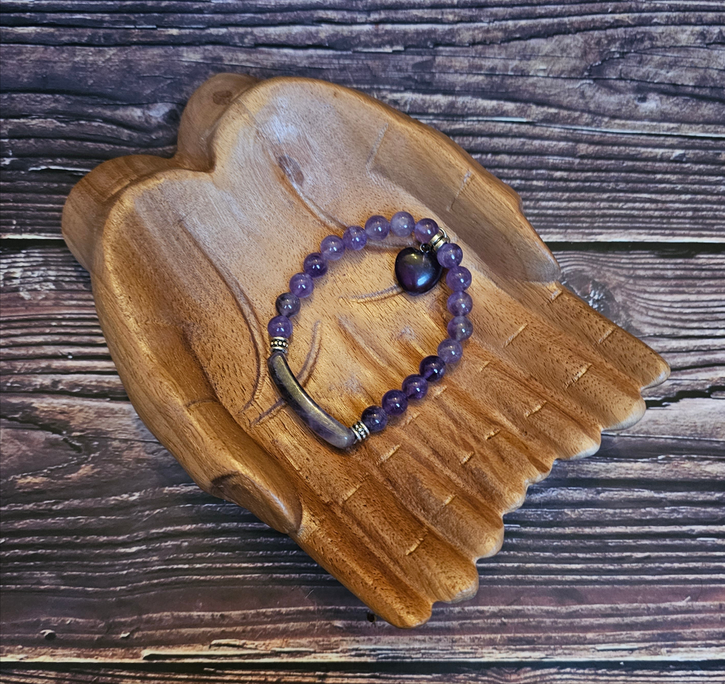 Wooden hands holding a purple amethyst bead bracelet with a heart-shaped bead and bar quartz accents.