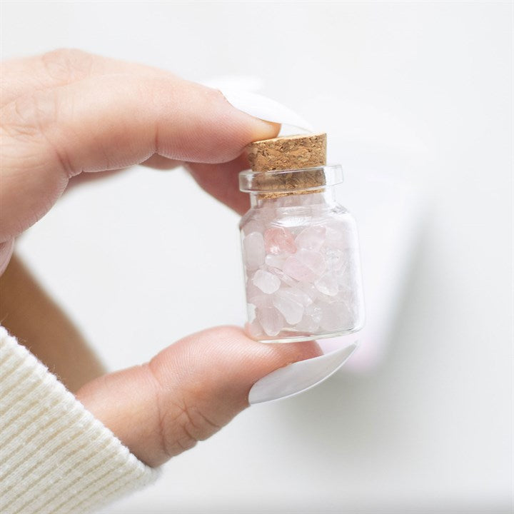 A hand holding a small glass jar filled with pink rose quartz crystal chips inside.