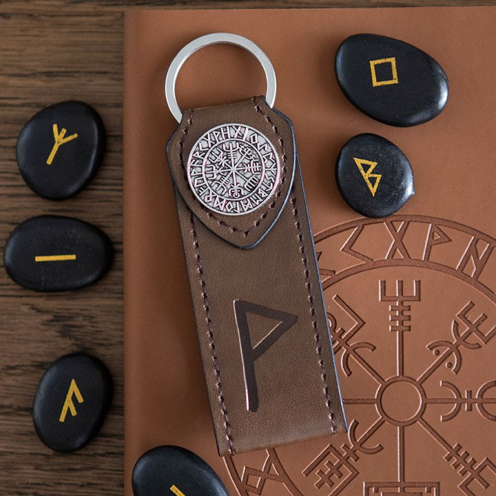 Faux brown leather keychain with metal emblem and runes, surrounded by black stones with yellow symbols on a brown surface.