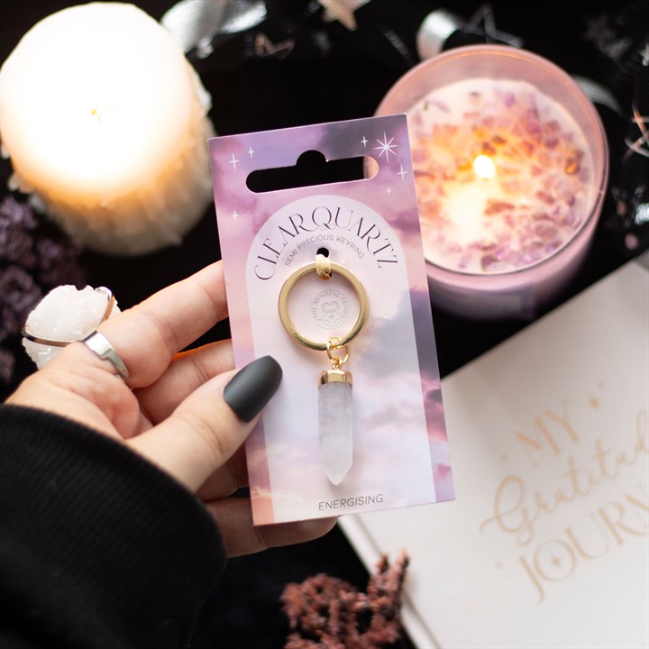 A hand holding a Clear Quartz crystal key rings on display and gold-coloured keyring attachment, presented on pink and purple backing cards with a star pattern.