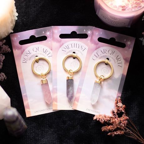 Three crystal key rings on display, each with a different colour of crystal (Amethyst, Clear Quartz, Rose Quartz) and gold-coloured keyring attachment, presented on pink and purple backing cards with a star pattern.