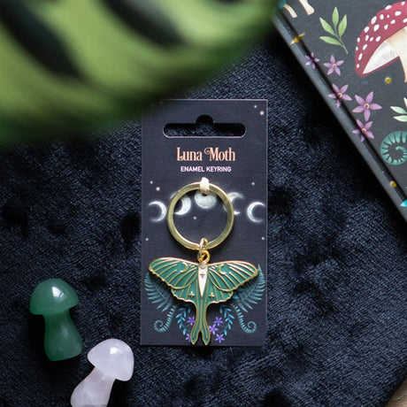 A gold-tone Luna Moth charm key ring on a black backing card with 'Luna Moth' text and moon phases illustrated at the top. The key ring is displayed on a dark fabric surface, alongside a green and a white mushroom.