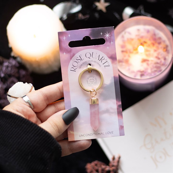 A hand holding a Rose Quartz crystal key rings on display and gold-coloured keyring attachment, presented on pink and purple backing cards with a star pattern.